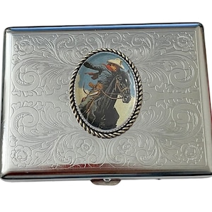 May include: Silver cigarette case with an engraved floral design and a round, gold-rimmed medallion featuring a cowboy riding a horse.