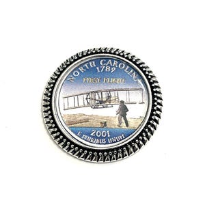 K&ouml;nnte beinhalten: Eine silberfarbene M&uuml;nze mit einem blauen und wei&szlig;en Bild des ersten Fluges der Gebr&uuml;der Wright in North Carolina. Die M&uuml;nze tr&auml;gt den Text "NORTH CAROLINA 1789 FIRST FLIGHT 2001 E PLURIBUS UNUM" am Rand.