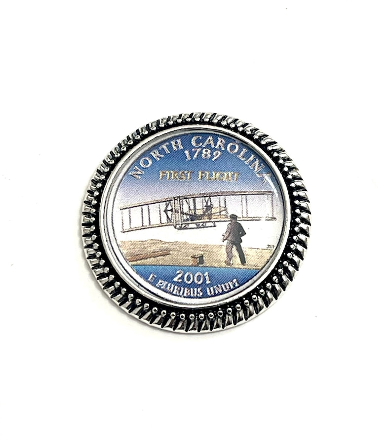 K&ouml;nnte beinhalten: Eine silberfarbene M&uuml;nze mit einem blauen und wei&szlig;en Bild des ersten Fluges der Gebr&uuml;der Wright in North Carolina. Die M&uuml;nze tr&auml;gt den Text "NORTH CAROLINA 1789 FIRST FLIGHT 2001 E PLURIBUS UNUM" am Rand.