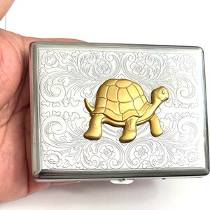 May include: A silver metal cigarette case with an engraved floral pattern and a gold-colored turtle emblem. The case is rectangular with rounded edges and a hinged closure. The turtle is in a standing position.