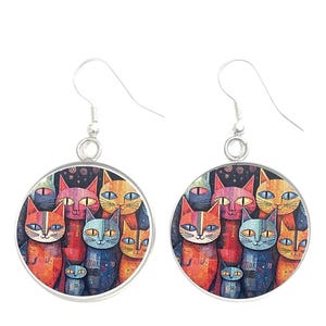 以下が含まれることがあります： カラフルな猫のイラストが描かれた丸いイヤリングのペア。イヤリングはシルバーカラーのフックと、オレンジ、ブルー、ピンクの色合いの様々な猫の精巧なデザインが特徴です。猫の目は黄色です。