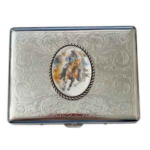 May include: Silver cigarette case with an engraved floral design and a round, silver-toned medallion featuring a cowboy riding a horse. The medallion has a white background and a colorful image of the cowboy and horse.
