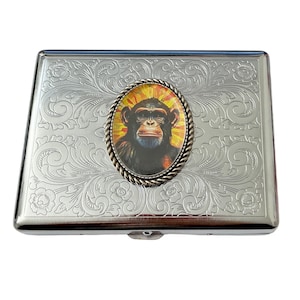 May include: Silver cigarette case with an engraved floral design and a round, silver-toned medallion featuring a colorful image of a chimpanzee.