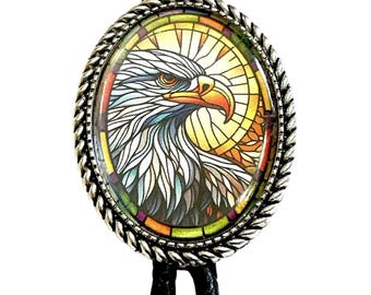 May include: Oval bolo tie with an eagle design. The silver pendant has a stained-glass style eagle head with a yellow, orange, and green background. The black braided cord completes the western-inspired accessory.