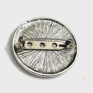 Op de afbeelding: Zilverkleurige ronde broche met een gestructureerd, radiaal ontwerp. De achterkant heeft een speldsluiting voor een veilige bevestiging. Deze broche is een klassiek accessoire, geschikt om kleding of accessoires een vleugje elegantie te geven.