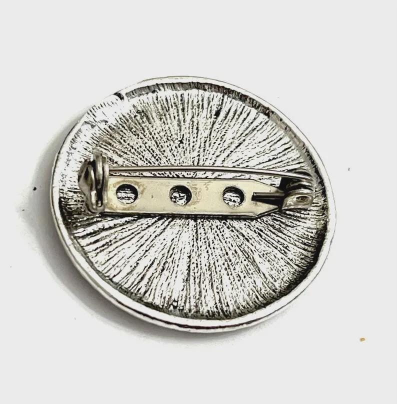 Op de afbeelding: Zilverkleurige ronde broche met een gestructureerd, radiaal ontwerp. De achterkant heeft een speldsluiting voor een veilige bevestiging. Deze broche is een klassiek accessoire, geschikt om kleding of accessoires een vleugje elegantie te geven.
