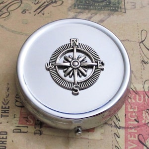 May include: A silver-colored, round pill box with a compass design on the lid. The compass is detailed with directional indicators and a central starburst design. The box has a hinged opening and a polished, reflective surface.