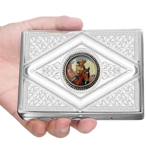 May include: Silver cigarette case with a decorative diamond pattern and a central circular image of a cowgirl on a horse. The case has a hinged lid and a polished finish.