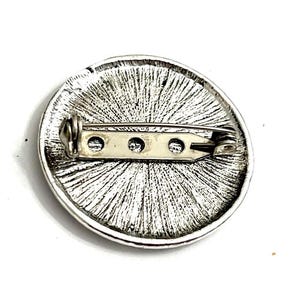 K&ouml;nnte beinhalten: Die R&uuml;ckseite einer runden silbernen Brosche mit einer Nadel und einem Verschluss. Die Brosche hat eine strukturierte Oberfl&auml;che mit einem radialen Muster.