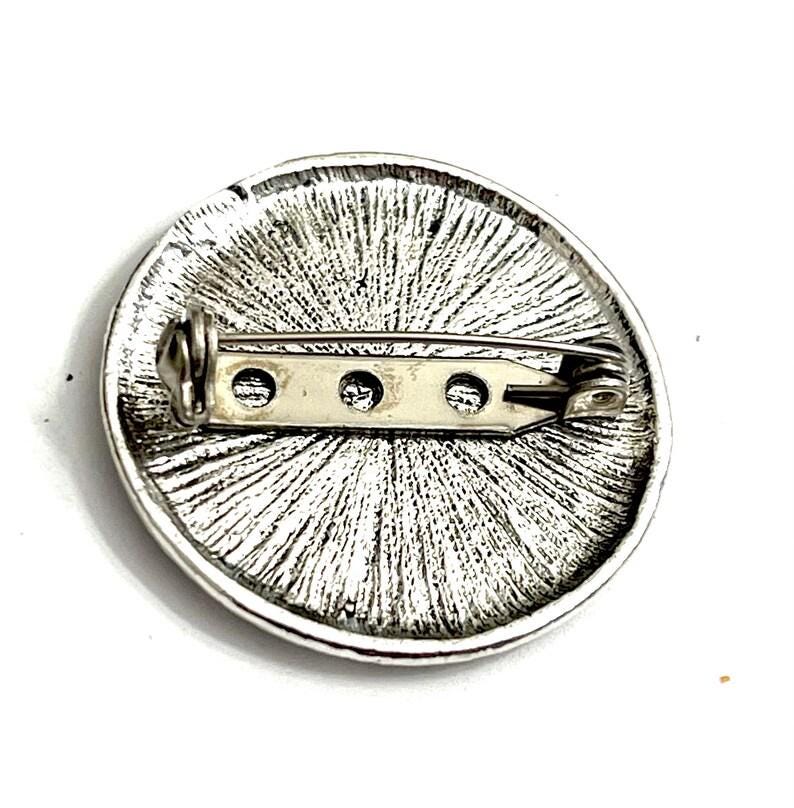 K&ouml;nnte beinhalten: Die R&uuml;ckseite einer runden silbernen Brosche mit einer Nadel und einem Verschluss. Die Brosche hat eine strukturierte Oberfl&auml;che mit einem radialen Muster.