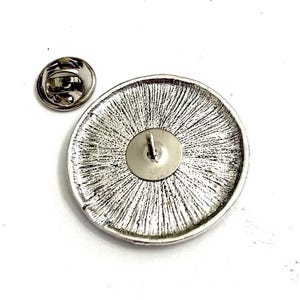 Op de afbeelding: Een zilverkleurige ronde pin met een textuurontwerp, met een pinrug en een aparte sluiting. De pin heeft een gedetailleerd, radiaal patroon op het oppervlak, tegen een witte achtergrond.