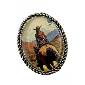 Puede incluir: Hebilla de cinturón ovalada plateada con una ilustración de temática occidental. La imagen representa a una persona montando a caballo por un paisaje desértico. La hebilla tiene un borde en forma de cuerda.