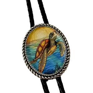 May include: A silver bolo tie with a round, glass pendant featuring a colorful illustration of a sea turtle swimming in the ocean. The pendant has a rope-like border.