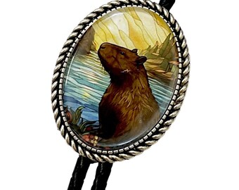 May include: A silver bolo tie with a black cord features a round, glass cabochon with a colorful illustration of a capybara in a watery setting.