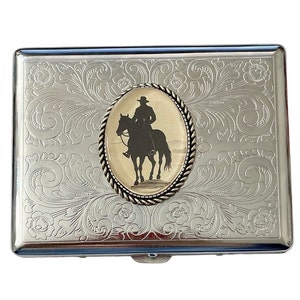 May include: Silver metal cigarette case with an engraved floral pattern. An oval inset features a silhouette of a cowboy on a horse. The case has a hinged lid and a secure clasp. A stylish accessory.
