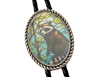 May include: A black bolo tie with a silver oval pendant featuring a stained glass image of a raccoon in a forest setting.