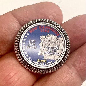 May include: A silver-colored coin with a blue and white image of the Old Man of the Mountain, a rock formation in New Hampshire. The coin has the words "New Hampshire 1788" and "Live Free or Die" around the edge. The coin also has the year "2000" and the words "E Pluribus Unum" on the bottom.