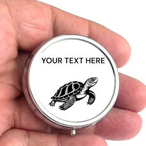 May include: A silver round pill box with a white lid featuring a black silhouette of a sea turtle and the text "YOUR TEXT HERE".