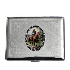 May include: A silver cigarette case with a detailed embossed geometric pattern. An oval inset features a painting of a cowboy on horseback. The case has a hinged lid and a secure clasp.