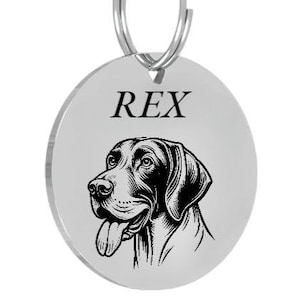 Op de afbeelding: Een ronde, zilveren hondenpenning met een zwart-witte illustratie van een hondenhoofd. De naam "REX" staat boven de afbeelding van de hond. Een zilveren ring is bevestigd.