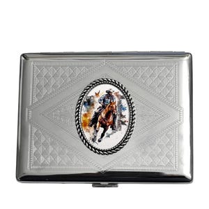 May include: A silver cigarette case with a detailed cowboy and horse illustration. The case has an oval image of a cowboy riding a horse, surrounded by a decorative border. The case has a geometric pattern.