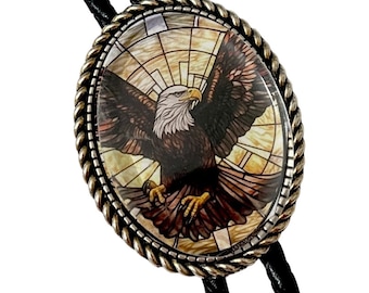 May include: An oval bolo tie with a silver-toned rope-style border. The centerpiece features a detailed image of a bald eagle with outstretched wings against a stained-glass-style background. The tie has a black braided cord.