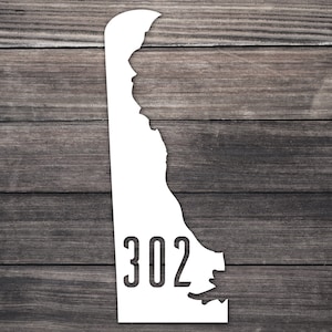 May include: White outline of the state of Delaware with the area code 302 printed inside. The outline is on a dark brown wood background.