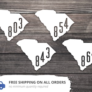 May include: Four white outline maps of South Carolina with black numbers inside. The numbers are 803, 854, 843, and 864. The maps are on a wood background.