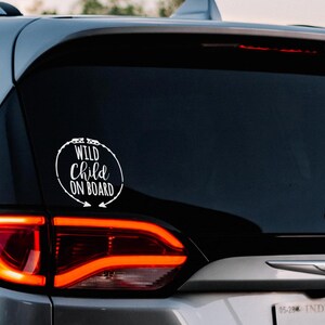 May include: A white vinyl sticker on the rear window of a silver car. The sticker reads "Wild Child On Board" in a circle with arrows pointing to the right.