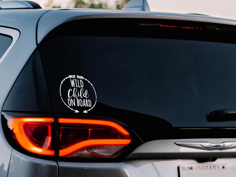 May include: A white vinyl sticker on the rear window of a silver car. The sticker reads "Wild Child On Board" in a circle with arrows pointing to the right.