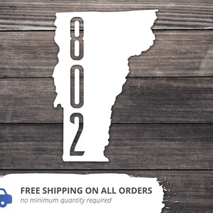 May include: White silhouette of the state of Vermont with the area code 802 printed vertically inside the state outline. The background is a dark wood grain.