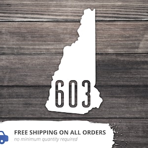 May include: White outline of the state of New Hampshire with the number 603 inside. The outline is on a wood background.