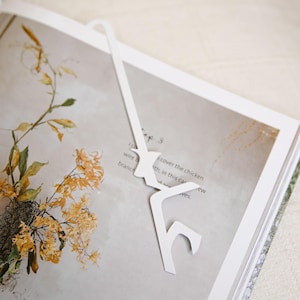 May include: A white, flat, elongated bookmark with a stylized, angular cutout design at one end, resting on the open pages of a book that features a printed illustration of dried yellow flowers and visible text on the page.