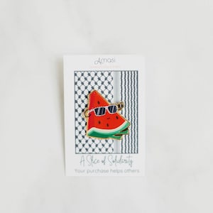 Può includere: Una spilla in smalto dorata con un'anguria di cartone animato che indossa occhiali da sole. L'anguria è rossa con la buccia verde e semi neri. La spilla è su una carta bianca con il testo "A Slice of Solidarity" e "Your purchase helps others".