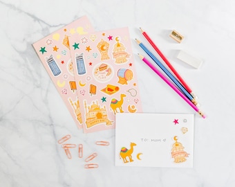 May include: Two light pink sticker sheets featuring colorful illustrations of camels, lanterns, mosques, and stars, alongside a white envelope with a camel and lantern sticker, three colored pencils, a sharpener, an eraser, and copper paper clips.