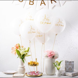 May include: White balloons with gold lettering that says "Eid Mubarak" and a mosque design. The balloons are tied together with gold string and are floating in the air. There are flowers in vases and a gold tiered stand with sweets on it.