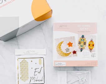 May include: Amasi Elevated Eid Decor & More DIY String Art Kit with two designs, including a crescent moon and star and a lantern. The kit includes white string, a wooden frame, and instructions. The box is white with pink and gold accents. The kit is suitable for ages 10 and up.