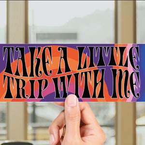 May include: A colorful sticker with the text "Take a little trip with me" in a retro font. The sticker has a purple, orange, pink, and blue background with a wavy pattern.