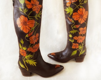Puede incluir: Un par de botas de cuero marrón de caña alta con un diseño floral. Las botas tienen una punta puntiaguda y un tacón de bloque. El diseño floral presenta flores naranjas y verdes.