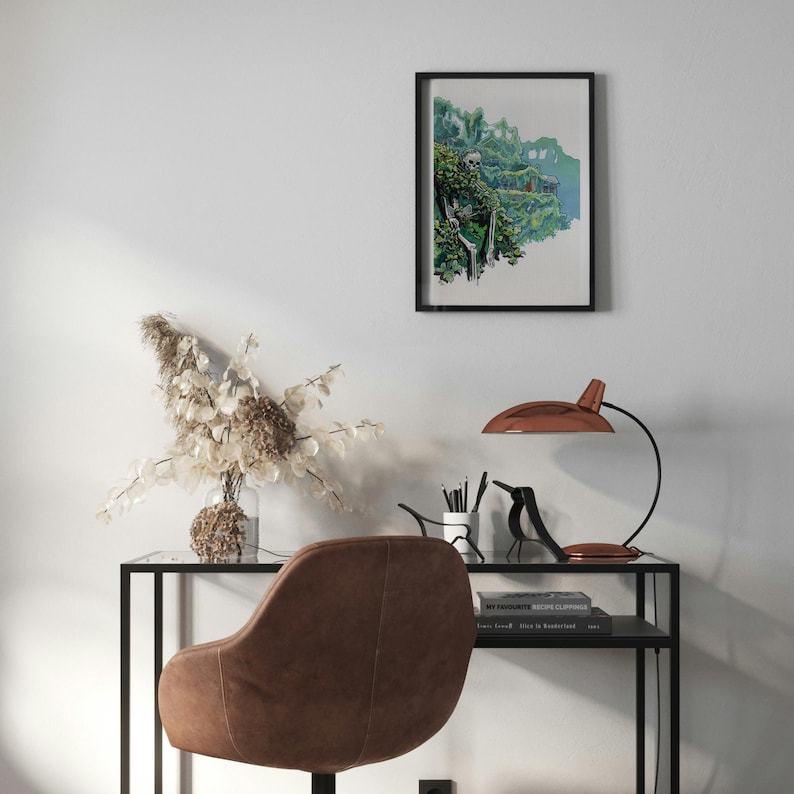 May include: A black framed print of a watercolor painting depicting a lush green landscape with a waterfall. The print is hanging on a white wall above a black desk with a copper lamp, a vase of dried flowers, and a brown leather chair.