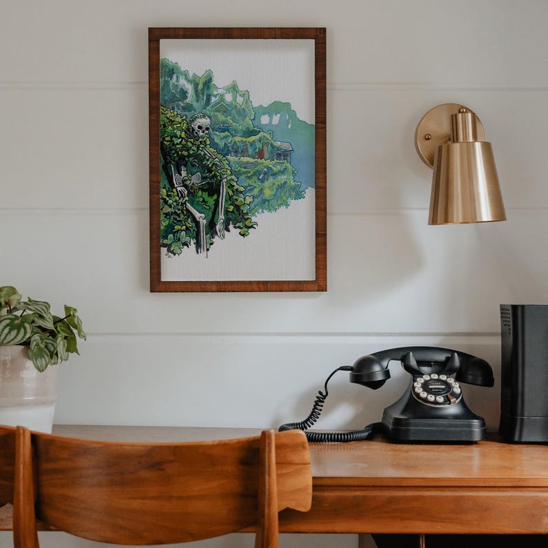 May include: A framed illustration of a skeleton sitting in a leafy green landscape. The illustration is hanging on a white wall above a wooden desk with a black rotary phone and a gold wall sconce.