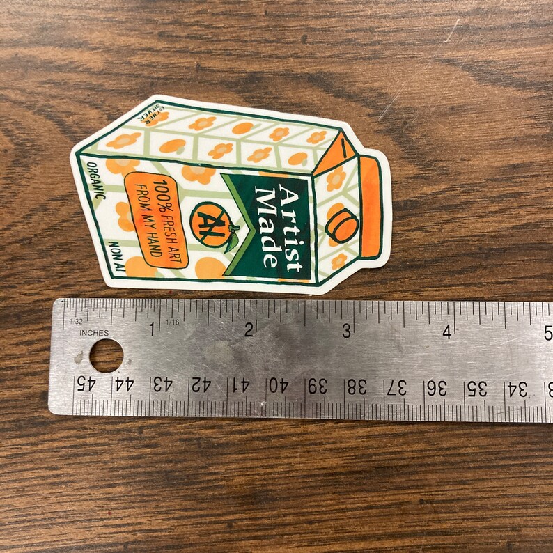 May include: A white sticker with a cartoon illustration of a juice box. The juice box is orange and green with the text "Artist Made" and "100% Fresh Art From My Hand" on it.