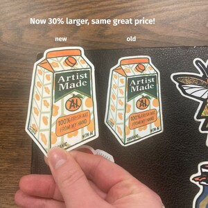 May include: Two orange juice carton stickers, one larger than the other. Both stickers feature the text "Artist Made" and "100% Fresh Art From My Hand". The larger sticker is labeled "new" and the smaller sticker is labeled "old".