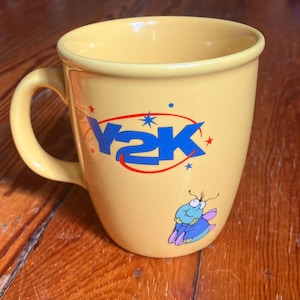 May include: A pale yellow ceramic mug featuring the blue text Y2K encircled by a red oval with small blue and red stars. Below the text is a small illustration of a light blue bug with purple wings and pink feet resting its chin on its hands.