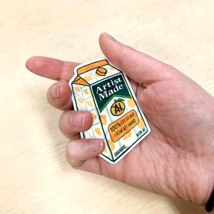 May include: A sticker of a cartoon orange juice carton with the text "Artist Made" and "100% Fresh Art From My Hand" in green text. The carton has a green "AI" symbol crossed out with a green line. The carton is labeled "Organic" and "Non AI".