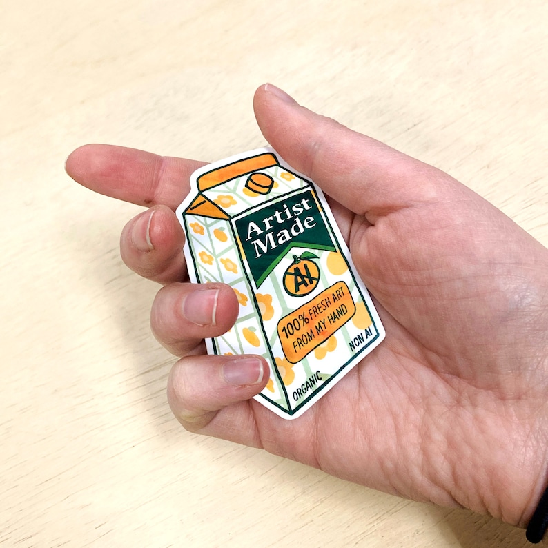 May include: A sticker of a cartoon orange juice carton with the text "Artist Made" and "100% Fresh Art From My Hand" in green text. The carton has a green "AI" symbol crossed out with a green line. The carton is labeled "Organic" and "Non AI".