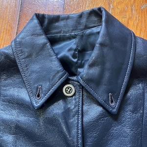 May include: A dark navy blue leather jacket featuring a classic pointed collar with two visible buttonholes on the collar points. The jacket has a single centred button closure and a smooth, dark interior lining visible at the neckline.