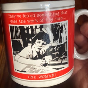 Op de afbeelding: Een witte keramische mok met een rode band en een zwart-witte illustratie van een persoon aan een bureau met een typemachine. De tekst luidt: They've found something that does the work of five men... ONE WOMAN! De mok heeft een wit handvat.