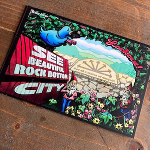 Op de afbeelding: Een kleurrijke ansichtkaart met de tekst "See Beautiful Rock Bottom City" en "Lookout!". Het toont een blauwe vogel met een verrekijker, kabouters en een schilderachtige achtergrond. De ansichtkaart heeft een vintage, grillig ontwerp.
