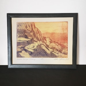 Pode incluir: Uma impressão emoldurada de uma gravura de paisagem intitulada "Golden Canyon Etching" com a assinatura do artista e a data "Lora Turner 2019". A gravura representa um cânion rochoso com uma vista distante de um vale em tons de laranja, amarelo e roxo.